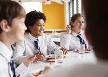 Group Of High School Students Wearing Uniform Sitting Around Table And Eating Lunch In Cafeteria