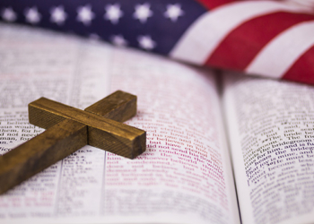 A holy Christian cross laying on an open Bible with an American flag in the background.