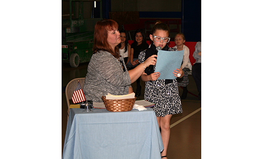 With her teacher, Cheryl DeVecchis, holding the microphone, first grade student Samantha Brown reads her essay. DeVecchis has been teaching her students at St. Michael the Archangel Regional School, Clayton, how to write persuasive essays and present their arguments. Because she compared the effort to what lawyers do in a courtroom, the boys and girls had to opportunity to wear business attire, carry briefcases and  present their “cases” to a court consisting of family members, teachers and students.