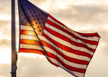 Stars and stripes American flag flying on a flag pole, backlit by sunlight against a sky