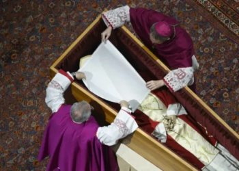 Before the wooden coffin is closed, Pope Benedict XVI’s personal secretary Archbishop Georg Gänswein and Monsignor Diego Giovanni Ravelli, the Vatican’s lead master of ceremonies for papal liturgies, place a white veil over the late pope’s face. The action on Jan. 4, 2023, is part of the funeral rites for popes. Photo-Vatican Media