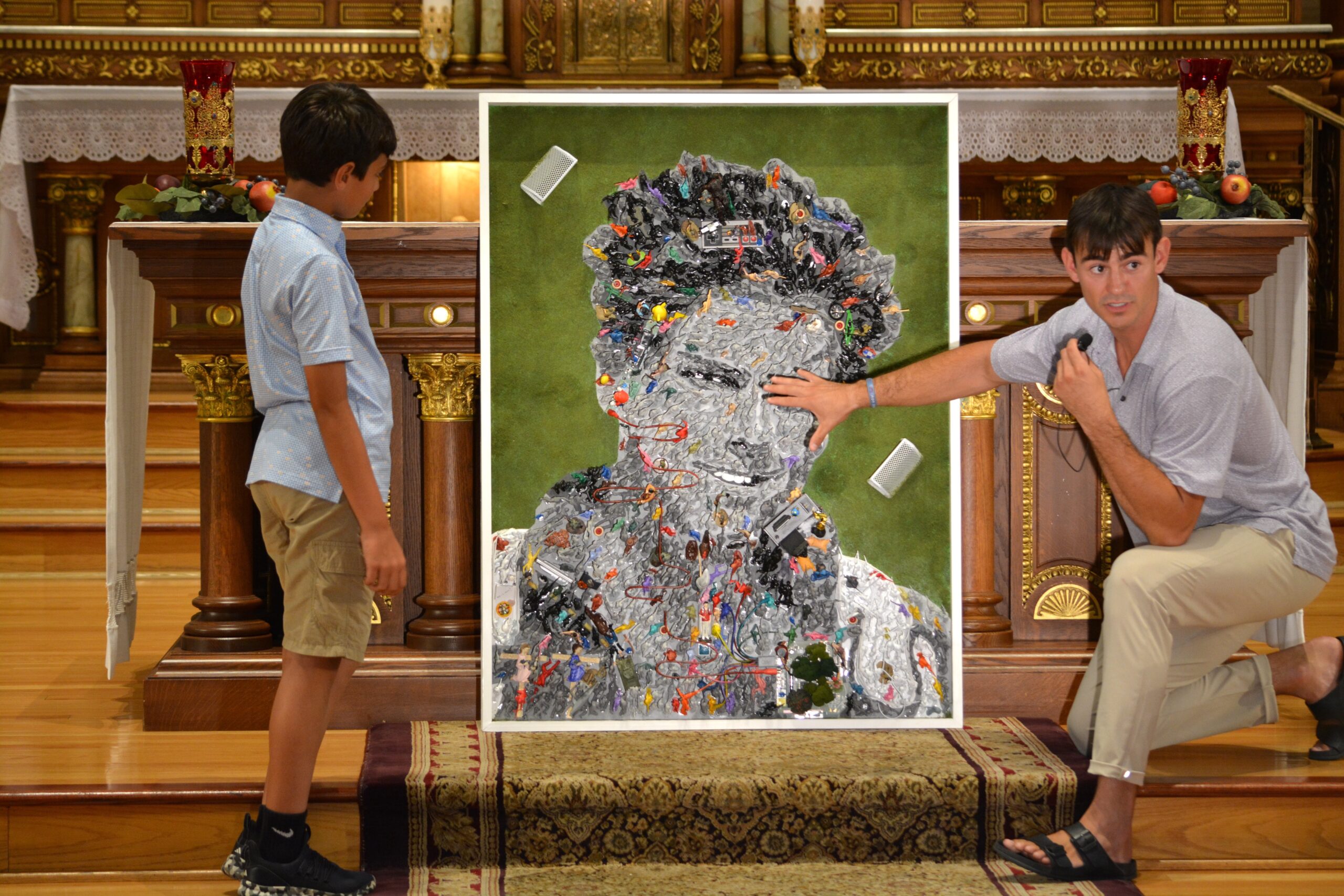Denver-based multimedia artist Johnny Vrba is pictured inside St. Hedwig Church in Chicago July 23, 2025,  explaining the significance of a section of the Blessed Carlo Acutis mosaic he created for a Chicago parish bearing the soon-to-be-saint’s name, to answer a boy’s questions about the mosaic. The portrait made up of more than 1,000 toy figurines includes numerous aspects of the teenager’s life, along with biblical references and the 163 Eucharistic miracles that Blessed Acutis documented. (OSV News photo/Simone Orendain)