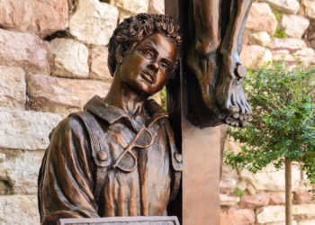 A sculpture of Blessed Carlo Acutis kneeling at the foot of the crucified Christ is seen in Assisi, Italy, Aug. 21, 2025, after a rainstorm. The bronze work titled "St. Carlo at the Cross" is by Canadian sculptor Timothy Schmalz, and it portrays the young blessed leaning his head against the cross while holding a laptop depicting the sacred vessels for Holy Communion. (CNS photo/Lola Gomez)