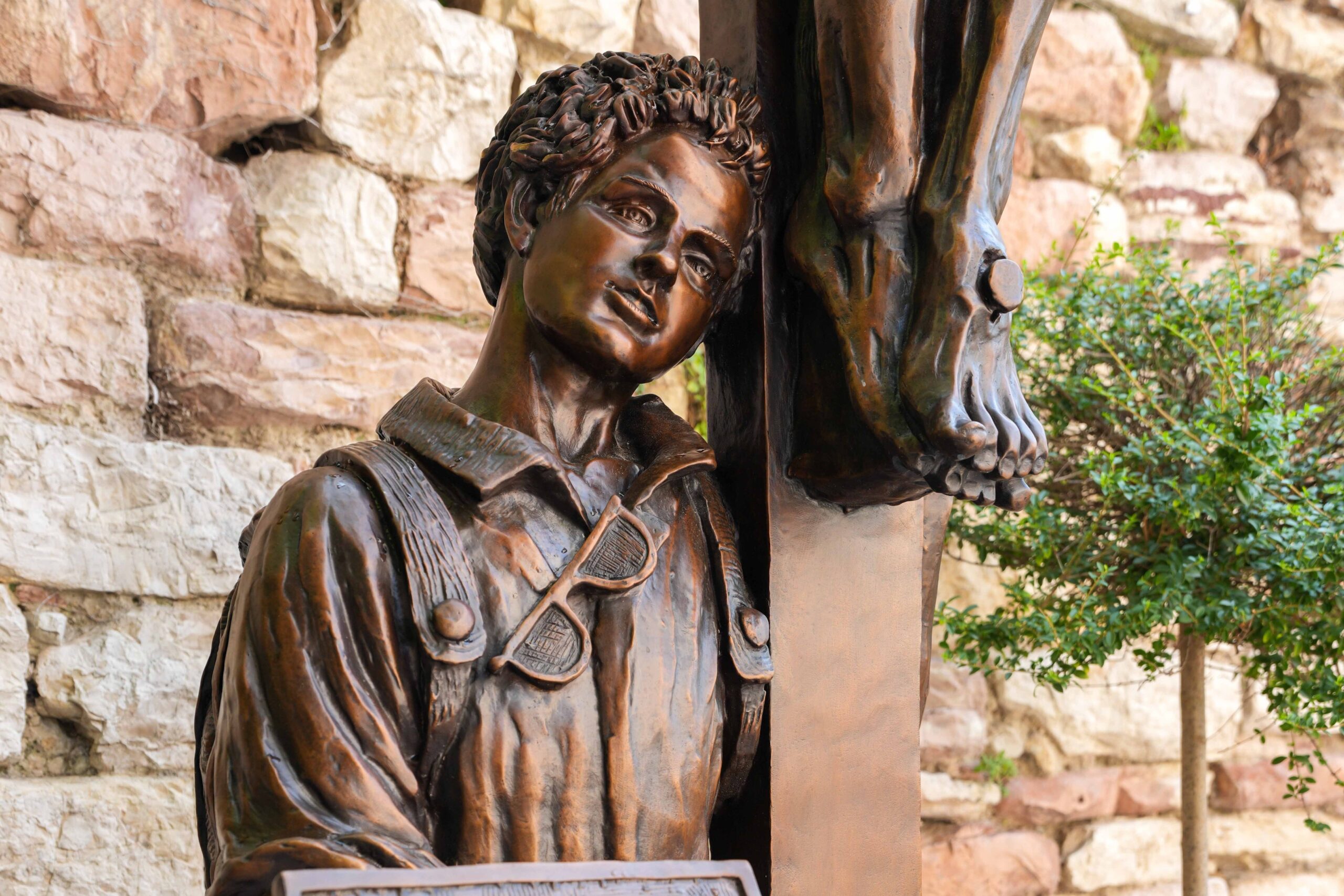 A sculpture of Blessed Carlo Acutis kneeling at the foot of the crucified Christ is seen in Assisi, Italy, Aug. 21, 2025, after a rainstorm. The bronze work titled "St. Carlo at the Cross" is by Canadian sculptor Timothy Schmalz, and it portrays the young blessed leaning his head against the cross while holding a laptop depicting the sacred vessels for Holy Communion. (CNS photo/Lola Gomez)