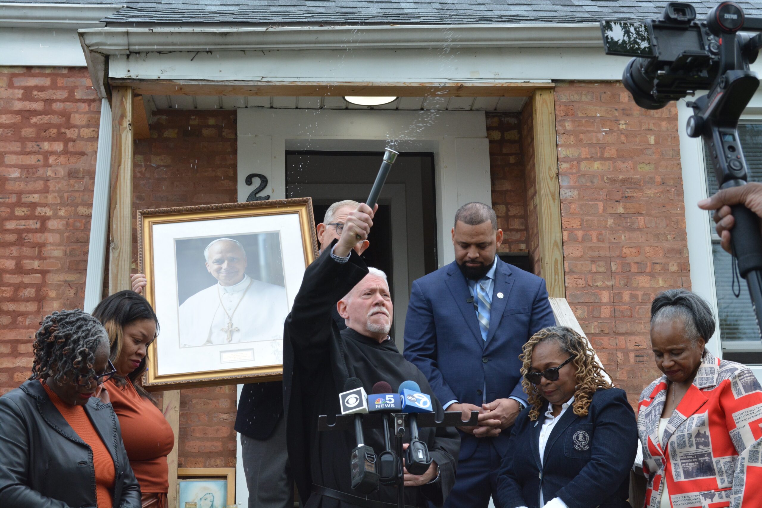 On the steps of Pope Leo XIV’s boyhood home in Dolton, Ill., a suburb of Chicago, Midwest Augustinians outgoing superior, Father Anthony B. Pizzo blesses the crowd after blessing the framed portrait of Pope Leo held by The Pope Leo XIV American Legacy Foundation chairman Vince Kolber, while Village of Dolton Mayor Jason House and his staff bow their heads. (OSV News photo/Simone Orendain)